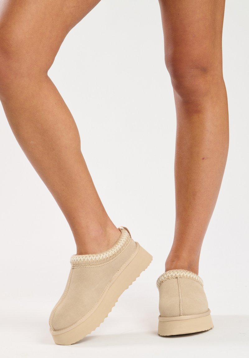 Beige slip-on shoes made of soft suede with a textured collar and a thick rubber sole. Features a rounded toe and minimal design details.