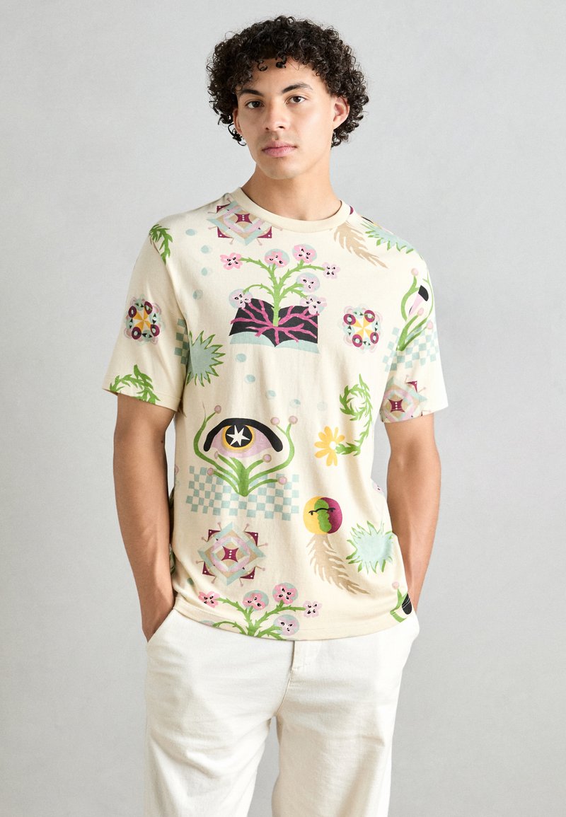 Cream short-sleeve t-shirt featuring colorful floral and geometric patterns, with a relaxed fit and soft texture. Paired with light-colored pants.