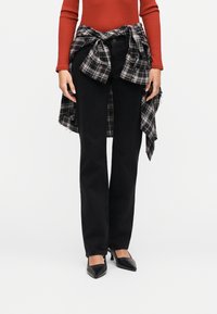 Femme portant un jean noir à jambes droites, des talons noirs, un haut à manches longues rouille et une chemise à carreaux noire et blanche nouée autour de la taille.