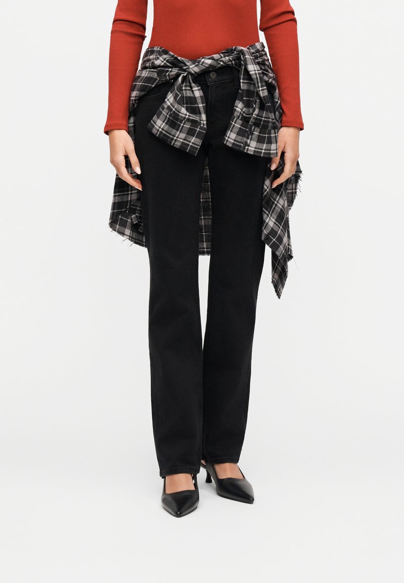 Femme portant un jean noir à jambes droites, des talons noirs, un haut à manches longues rouille et une chemise à carreaux noire et blanche nouée autour de la taille.