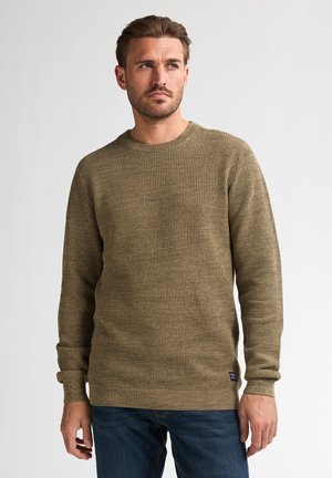 Man wearing olive green knit sweater and blue jeans, standing against plain white background, looking to his left.