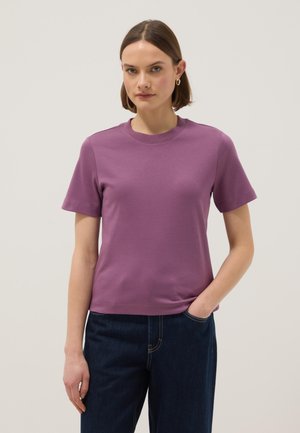 Mujer que lleva una camiseta de manga corta morada y jeans azul oscuro con una mano en el bolsillo, de pie frente a un fondo claro y liso.