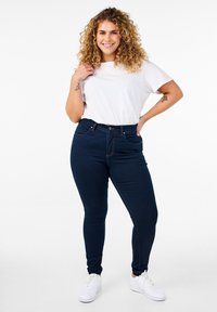 Jeans de tiro alto azul marino con cinco bolsillos, complementados con una camiseta blanca de cuello redondo y zapatillas blancas. Pelo rizado enmarcando la cara.