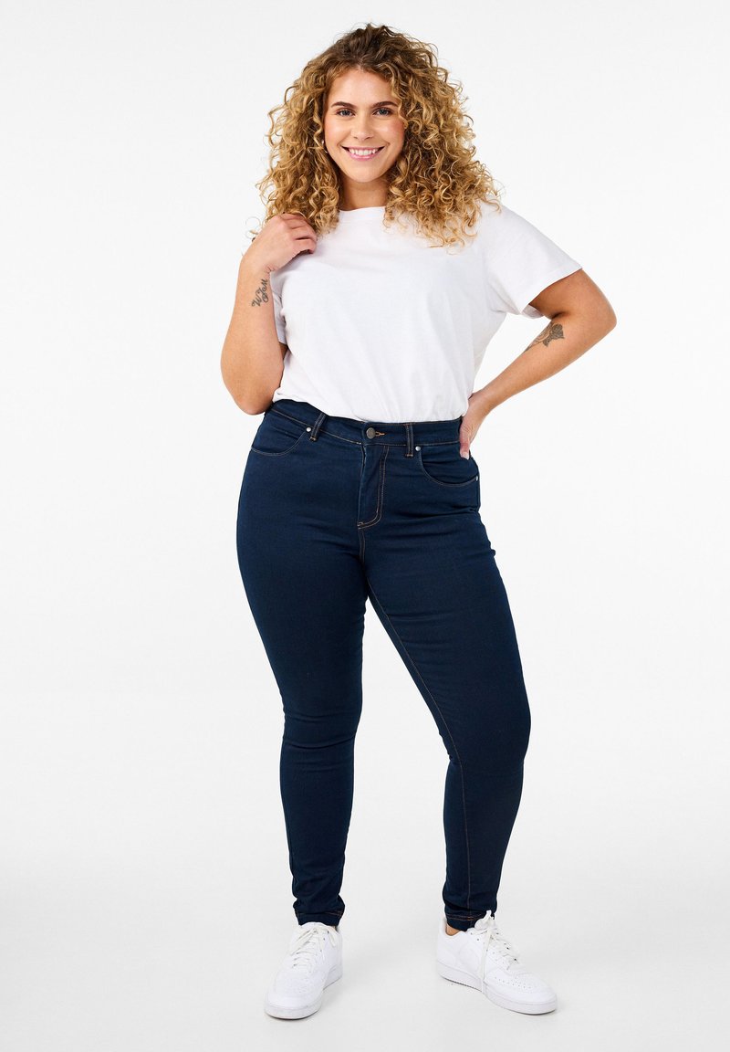 Jeans de tiro alto azul marino con cinco bolsillos, complementados con una camiseta blanca de cuello redondo y zapatillas blancas. Pelo rizado enmarcando la cara.
