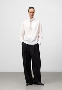 Camicia bianca a maniche lunghe con colletto alla coreana e dettaglio della cravatta, abbinata a pantaloni neri larghi. Tessuto morbido e vestibilità rilassata in tutto il capo.