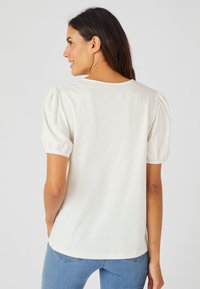 Top blanco de manga corta con mangas abullonadas, tejido suave, cuello redondo y un ajuste relajado. Combinado con jeans de mezclilla azules.