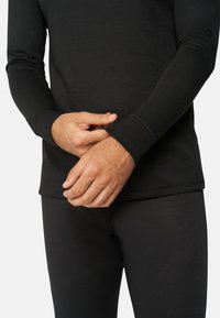 Schwarzes Thermo-Langarmshirt mit strukturiertem Finish, enganliegendem Schnitt und kontrastierenden Nähten. Das Model justiert den Ärmel, während es passende schwarze Hosen trägt.
