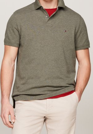 Homme portant un polo manches courtes vert olive par-dessus une chemise rouge, associé à un pantalon beige clair, debout avec une main dans la poche.