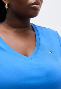 Nahaufnahme einer Person, die ein blaues V-Ausschnitt-Shirt mit einem kleinen Tommy Hilfiger-Logo auf der Brust und einen goldenen Creolen-Ohrring trägt.