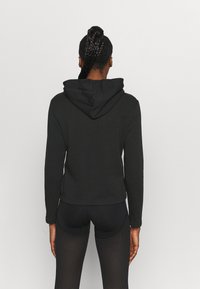 Sudadera negra con capucha, de manga larga, diseño ajustado y dobladillo acanalado. Llevada sobre leggings negros con un patrón de malla.