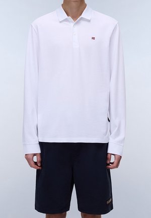 Mand iført en hvid langærmet polo med et lille rødt og hvidt logo kombineret med sorte knæhøje shorts.
