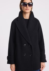 Czarny płaszcz dwurzędowy z grubego materiału, z szerokim kołnierzem, dużymi guzikami i zawiniętymi mankietami. Modelka nosi oversize'owe okulary przeciwsłoneczne.