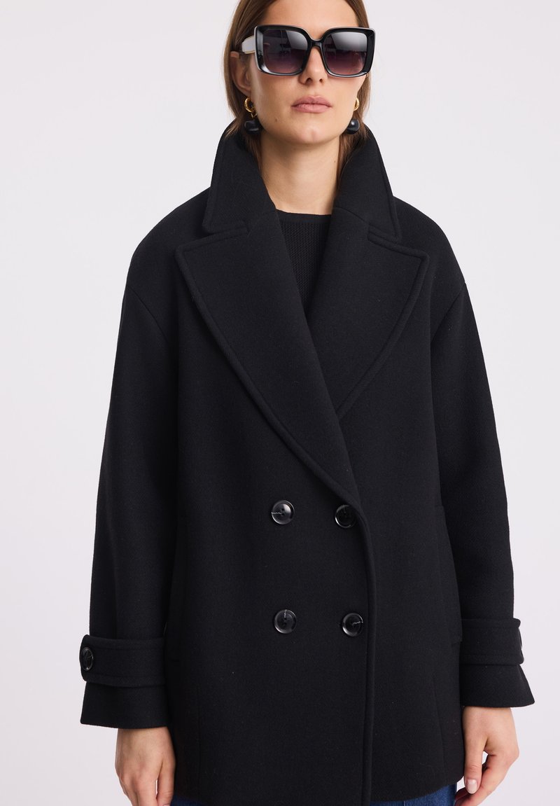 Czarny płaszcz dwurzędowy z grubego materiału, z szerokim kołnierzem, dużymi guzikami i zawiniętymi mankietami. Modelka nosi oversize'owe okulary przeciwsłoneczne.