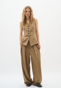 Gilet beige con bottoni neri, abbinato a pantaloni beige larghi e plissettati. Tessuto liscio con una vestibilità su misura. Design semplice, con decorazioni minime.