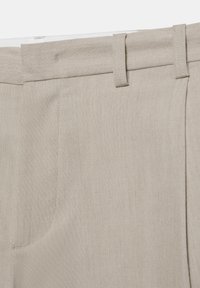 Gros plan de la taille d’un pantalon beige avec passants de ceinture, couture de la poche avant et patte de fermeture à bouton.