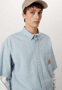 Chemise collier bleu clair avec un motif texturé, comportant une poche sur la poitrine gauche et un petit écusson logo orange. Chemise blanche à manches longues en dessous.