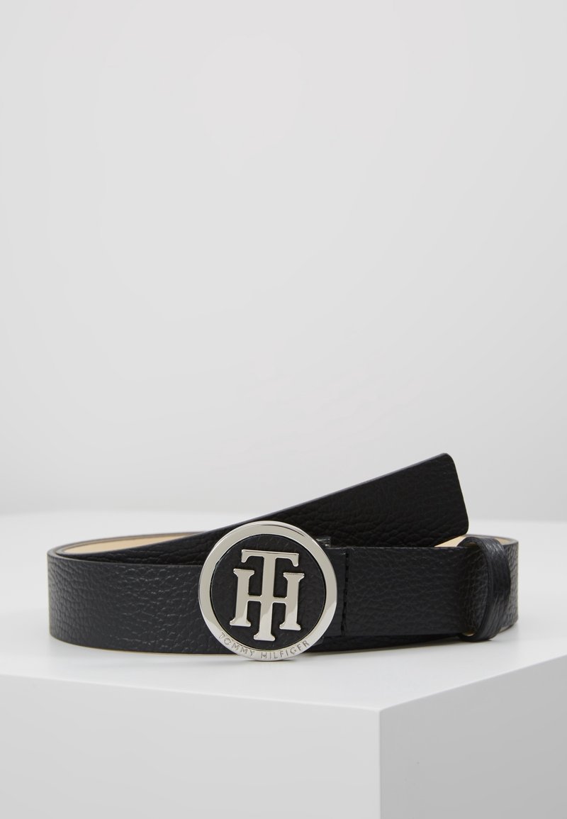 Ceinture en cuir noir texturé avec une boucle ronde en argent avec le logo Tommy Hilfiger sur une surface blanche.