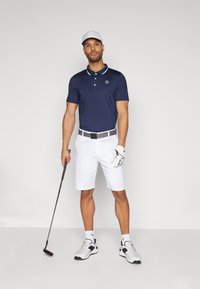 Polo shirt bleu marine avec des accents de col blanc, shorts blancs, ceinture noir et blanc, gant de golf blanc et chaussures de sport. Tenant un club de golf.