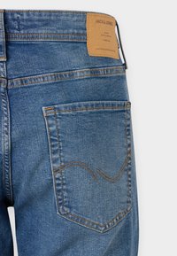 Džínsy v stredne modrej farbe s viditeľným prešívaním, s zadnou vreckom a hnedou koženou nášivkou s nápisom "JACK & JONES."