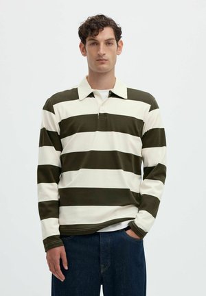 Homme portant un maillot de rugby à manches longues avec de larges rayures horizontales vert foncé et blanc et un jean bleu foncé, debout avec une main dans la poche.