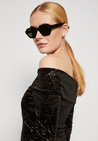 Occhiali da sole neri oversize con montatura angolare e lenti scure. Il modello indossa un top in velluto off-shoulder con motivi di stelle dorate.