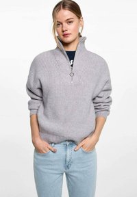Šedý rebrovaný pletený pulóver s polovičným zipsom, dlhými rukávmi a voľným strihom, skombinovaný so svetlomodrými džínsami.