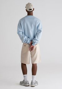 Sudadera azul clara, pantalones cortos beige, zapatillas plateadas y una gorra a rayas marrón usada al revés. Manos entrelazadas detrás de la espalda.