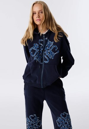 Sudadera con cremallera en navy con diseño de logo desgastado en azul. Tejido suave con un ajuste relajado. Pantalones a juego con acentos de logo similares en el dobladillo.