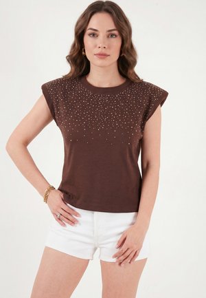 Femme aux cheveux bruns ondulés portant un haut marron sans manches avec de petits clous argentés et un short blanc, debout avec une main sur la hanche.