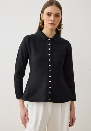 Femme portant un chemisier noir ajusté à manches longues avec col et boutons, et un pantalon blanc, debout devant un fond clair uni.