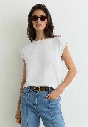 Kvinde iført hvid ærmeløs top, blå jeans med forreste sømme, brun bælte og mørke solbriller, stående foran en ensfarvet hvid væg.