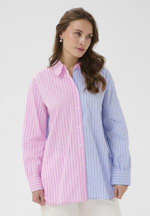 Ženska, ki nosi dolgo srajco z gumbi z rožnatimi in belimi črtami na eni strani ter modrimi in belimi črtami na drugi strani, stoji ob sivi steni.