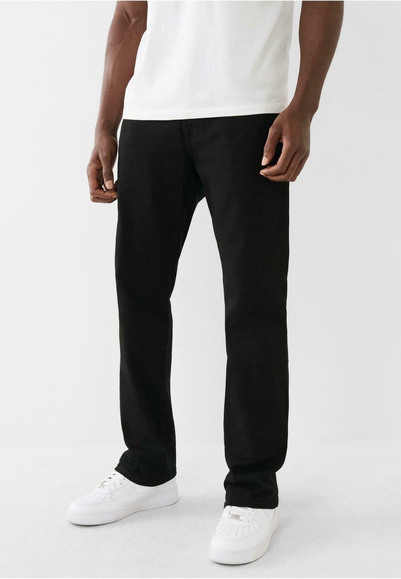Jeans negre cu picioare drepte din denim, cu un design clasic și detalii subtile de cusătură, asortați cu adidași albi și un tricou alb simplu.