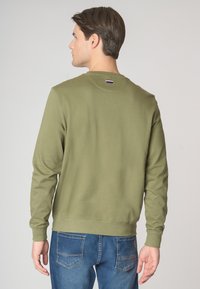 Felpa verde oliva con maniche lunghe, polsini e fondo a costine. Presenta una piccola etichetta del logo sul collo. Indossata con jeans denim blu.