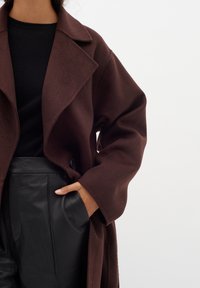 Hnědý vlněný kabát s klopami, oversized rukávy a detailním zapínáním; v kombinaci s černými koženými kalhotami. Textura vypadá jemně.