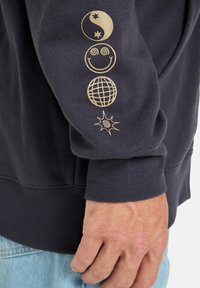 Navy sweatshirt with a crew neck, featuring gold graphic designs: yin-yang, smiley face, globe, and star accents on the sleeve.