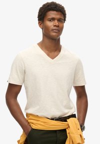 T-shirt beige con scollo a V realizzata in tessuto leggero, maniche corte, indossata con un felpa gialla legata in vita, pantaloni scuri e una cintura.
