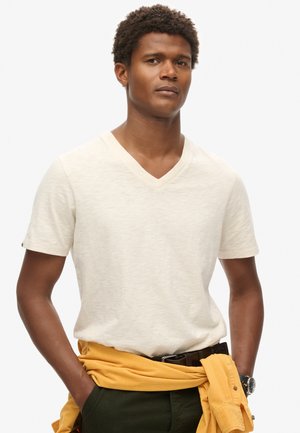 Beige V-hals T-shirt van lichtgewicht stof, korte mouwen, gedragen met een gele sweatshirt om de taille geknoopt, donkere broek en een riem.