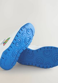 Weiße Sneaker mit leuchtend blauen Sohlen, die kreisförmige Muster aufweisen, sowie einem bunten Super Mario-Logo an der Seite, vor einem neutralen Hintergrund.