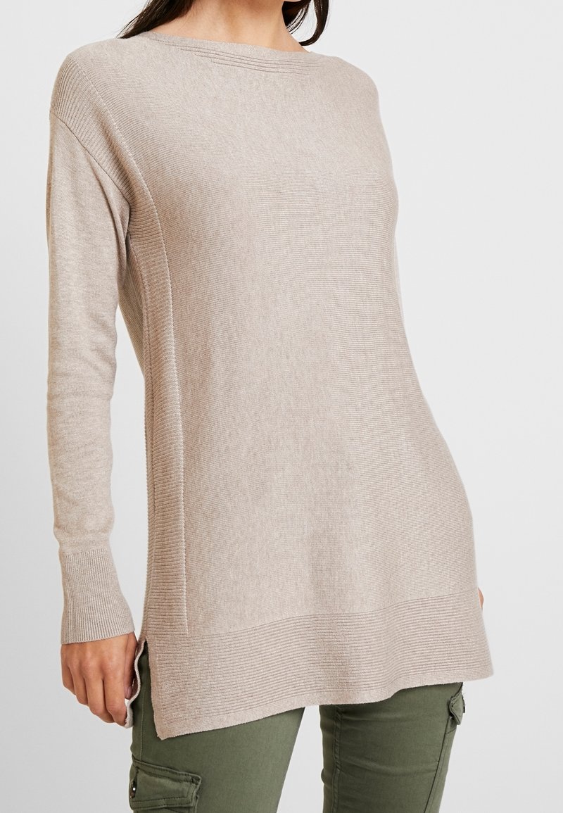 Femme portant un pull en maille beige à manches longues avec des fentes sur les côtés, associé à un pantalon cargo vert olive, vue du cou jusqu'à mi-cuisse.