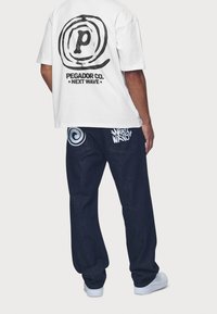 Vit t-shirt med svart runt logo på ryggen, kombinerad med mörkblå jeans som har ljusblå spiral- och textgrafik. Vita skor.