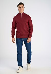 Roter Zip-Kragen-Pullover mit langen Ärmeln, kombiniert mit blauen Jeans und weißen Turnschuhen. Der Pullover hat eine strukturierte Oberfläche und gerippte Bündchen.