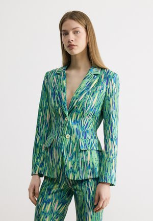 Femme aux cheveux lisses châtain clair portant un blazer à motifs verts et bleus avec un pantalon assorti, debout devant un fond blanc uni.
