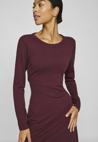 Långärmad burgundy klänning gjord av mjukt material, med rund halsringning och figurnära midja, med en jämn textur och sömlös design.