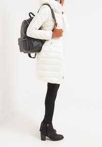 Vitt vadderat ytterplagg med hög krage, dragkedjestängning och sidofickor. Svart läderryggsäck och ankelstövlar med dragkedjedetalj. Slim fit svarta byxor.