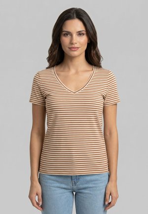 Donna con capelli scuri di lunghezza media, che indossa una maglietta a maniche corte con scollo a V a righe beige e bianche e jeans azzurri, in posizione neutra.