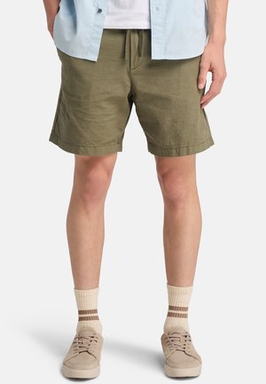 Pantaloni corti verde oliva realizzati in tessuto testurizzato, dotati di vita con cordoncino e vestibilità comoda con tasche laterali. Abbinati a sneakers beige e calzini a righe.