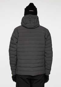 Vadderad grå jacka med huva, med horisontella quiltade sektioner, och en märkt baksidestab. Bärs med svarta handskar.