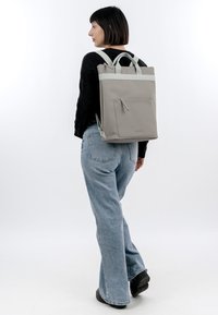 Frau mit kurzen dunklen Haaren, die einen schwarzen Pullover und blaue Jeans trägt und einen grauen rechteckigen Rucksack mit Frontreißverschlusstasche auf dem Rücken trägt.