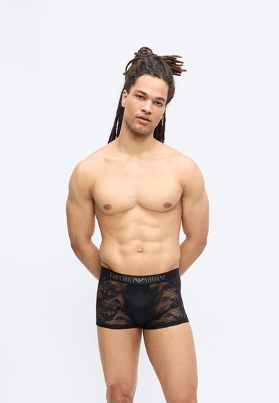 Μυώδης άνδρας με ντραντλοκς που φοράει μαύρα μπόξερ Emporio Armani με σχέδιο, στέκεται μπροστά σε έναν απλό λευκό φόντο.